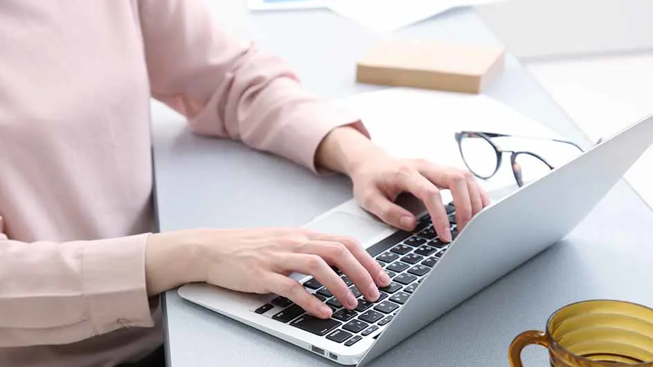 Een persoon typt op een laptop op een bureau, met een bril, mok en boek in de buurt.