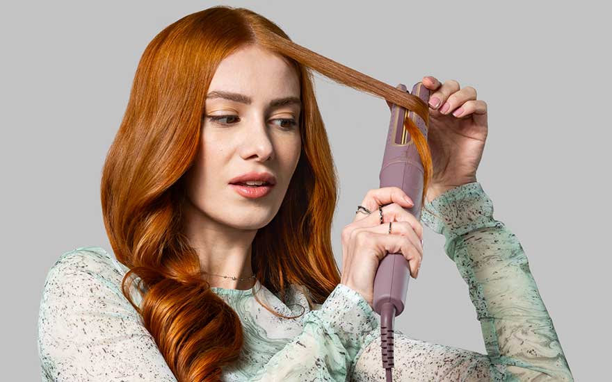 Persoon met rood haar in mintgroene blouse gebruikt een roze krultang om haar haar te stylen tegen een grijze achtergrond.
