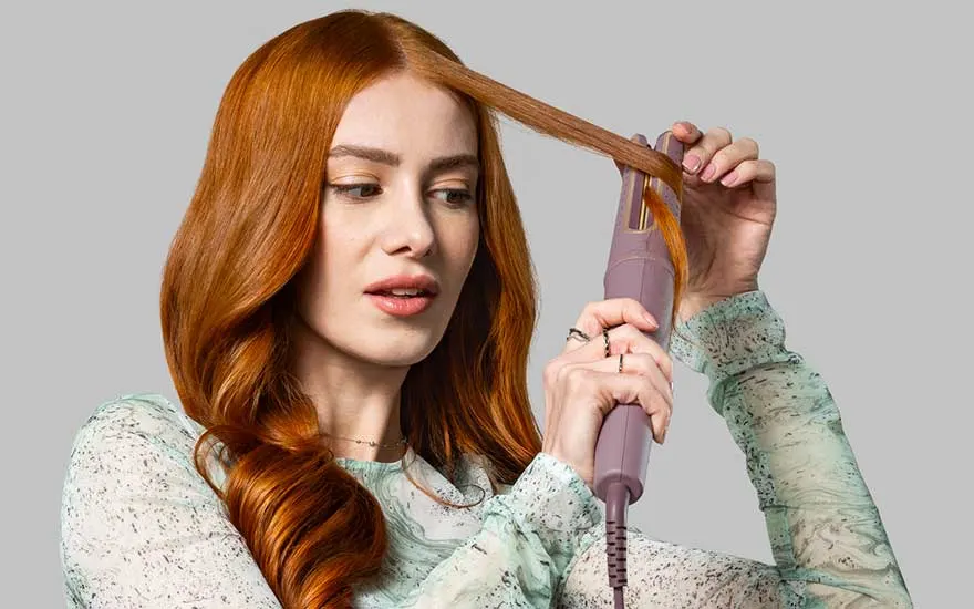 Persoon met rood haar in mintgroene blouse gebruikt een roze krultang om haar haar te stylen tegen een grijze achtergrond.