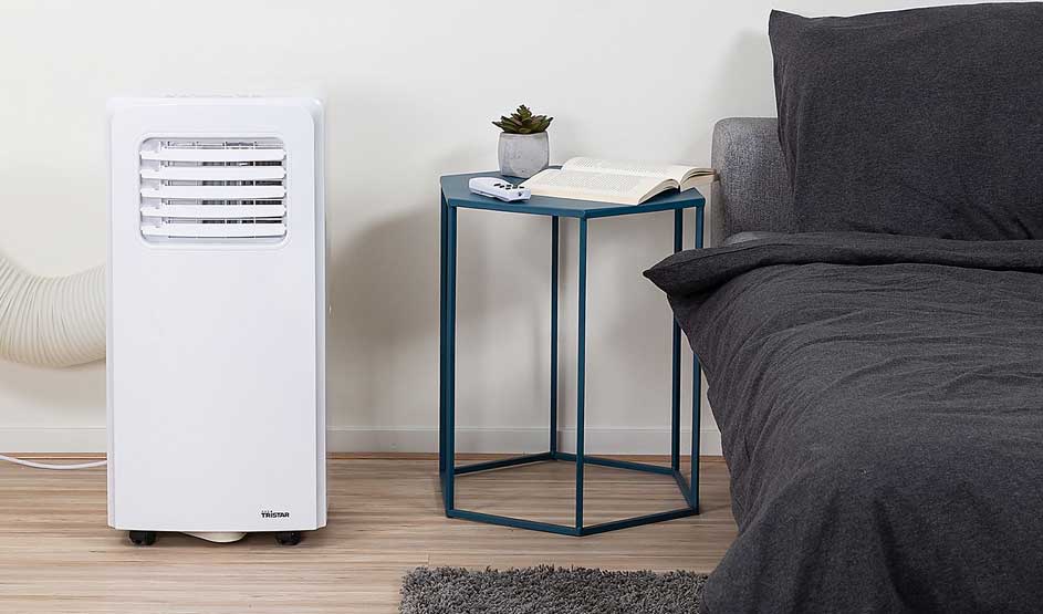 Een witte Tristar mobiele airco staat naast een bed en een blauw bijzettafeltje met een boek, plantje en afstandsbediening. Merk: Tristar.