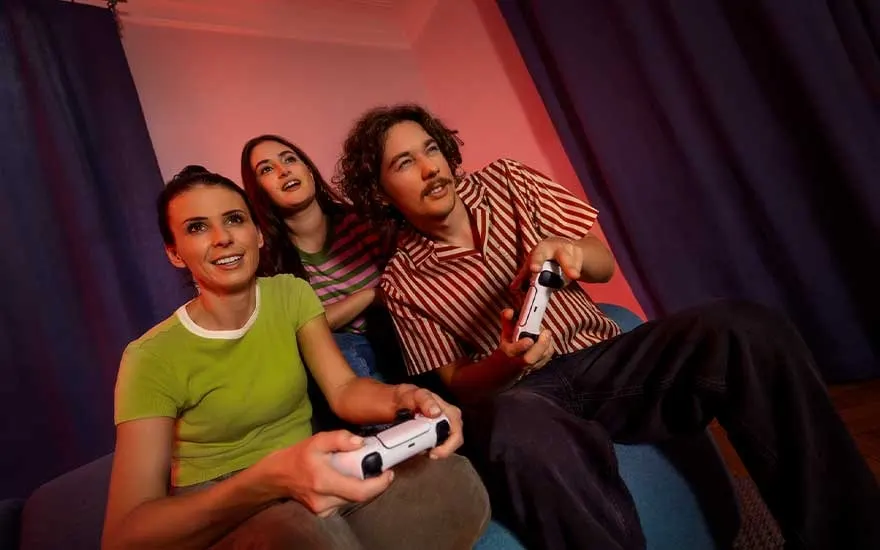 Three friends relax on the sofa, playing video games together with controllers in a warmly lit living room.