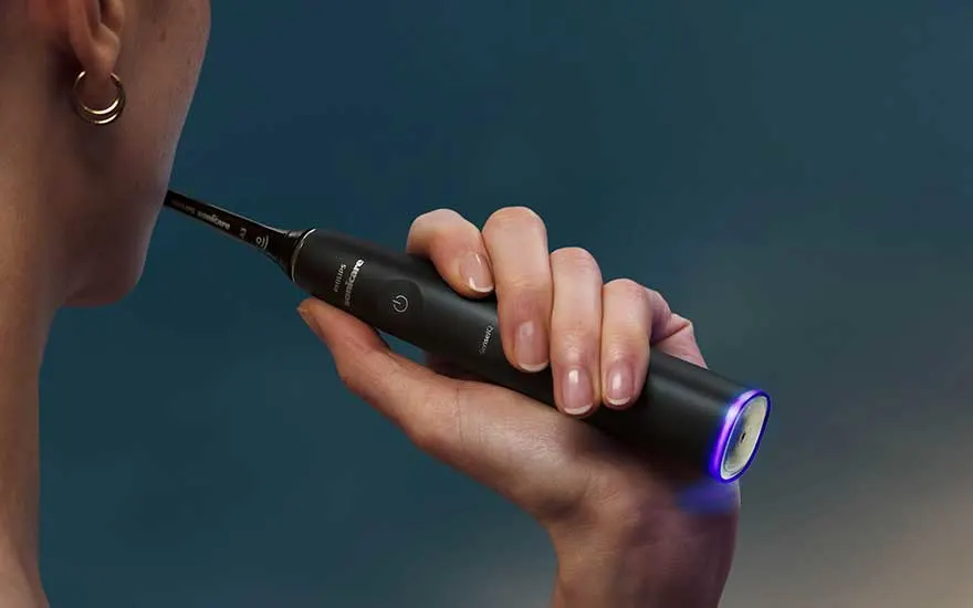 Een persoon houdt een zwarte sonische tandenborstel tegen de wang, met subtiele paarse verlichting op een donkere blauwe achtergrond.