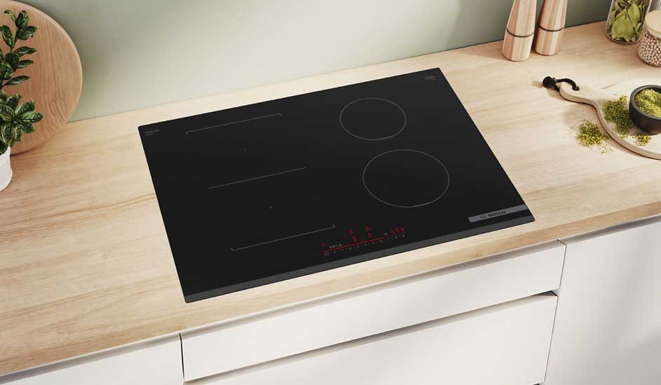Een zwarte inductiekookplaat ligt vlak in een licht houten werkblad, met rode touchbediening en kruidenpotjes op het aanrecht eromheen.