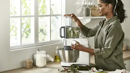 Een vrouw gebruikt een keukenmachine naast een raam. Op het aanrecht staan een pot met bloem, een courgette en knoflook.