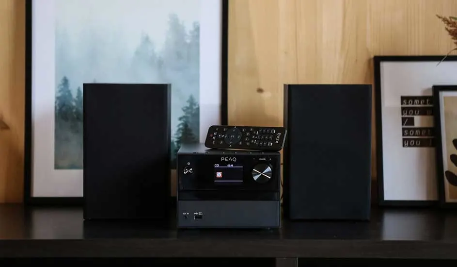A black stereo system with two speakers and a remote sits on a shelf, surrounded by framed art and a wooden wall background.