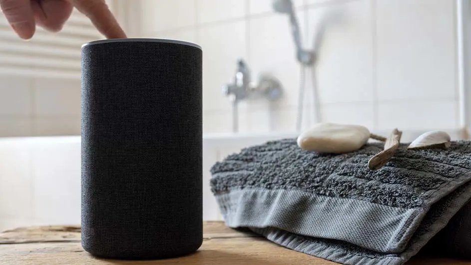 Een zwarte speaker wordt aangeraakt op een houten oppervlak, met een grijze handdoek, zeep, en een badkamer op de achtergrond.