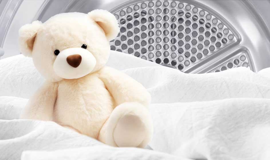 Een zachte teddybeer zit tussen witte lakens voor de trommel van een droger, waardoor het beeld schoon, warm en knus aanvoelt.