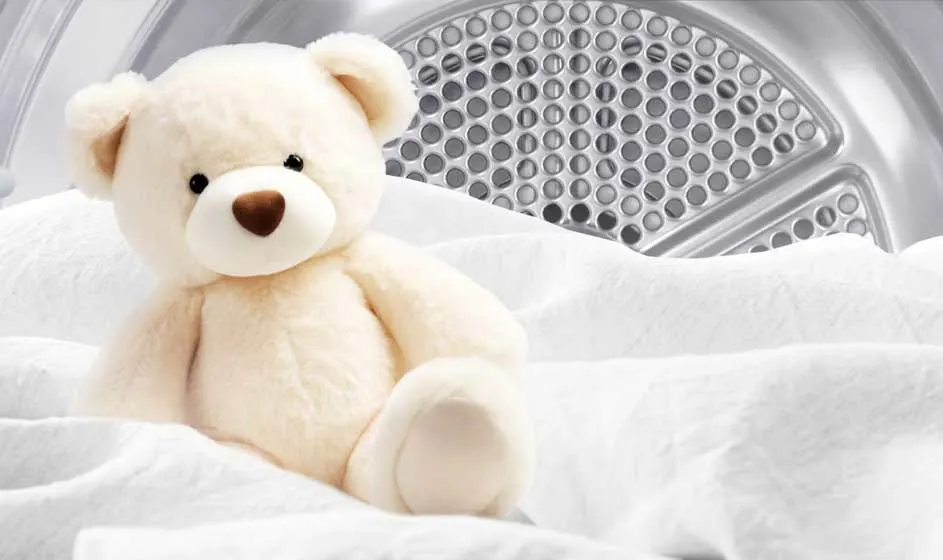 Een zachte teddybeer zit tussen witte lakens voor de trommel van een droger, waardoor het beeld schoon, warm en knus aanvoelt.