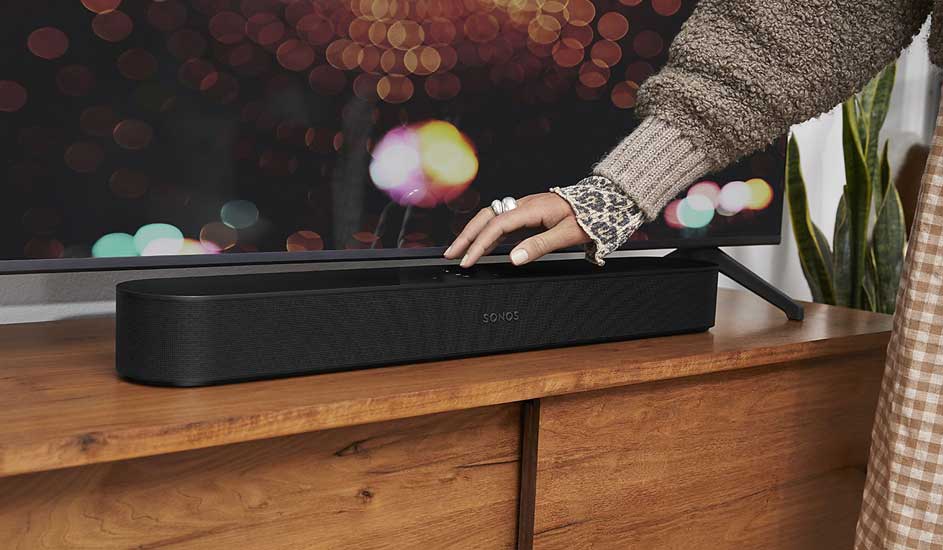 A hand adjusts a black Sonos soundbar on a wooden cabinet. In the background, a TV with bokeh effect creates a cosy, stylish atmosphere.