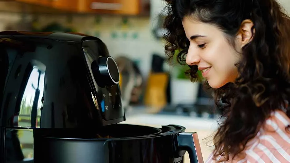 Een vrouw glimlacht naar een open zwarte airfryer in een keuken.