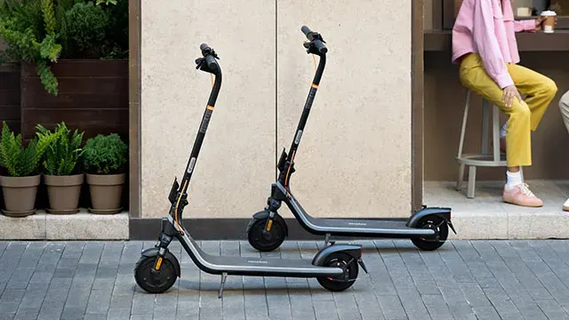 Twee zwarte elektrische scooters staan op een stenen trottoir voor een gebouw met plantenbakken en een zittend persoon.