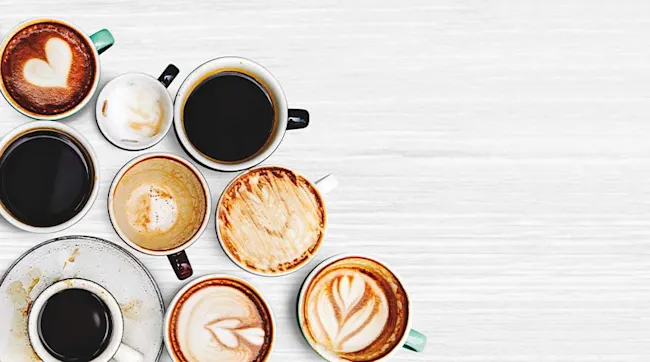 Overhead view of nine cups, some with latte art, on a white wooden surface. Some cups are full, some are empty.