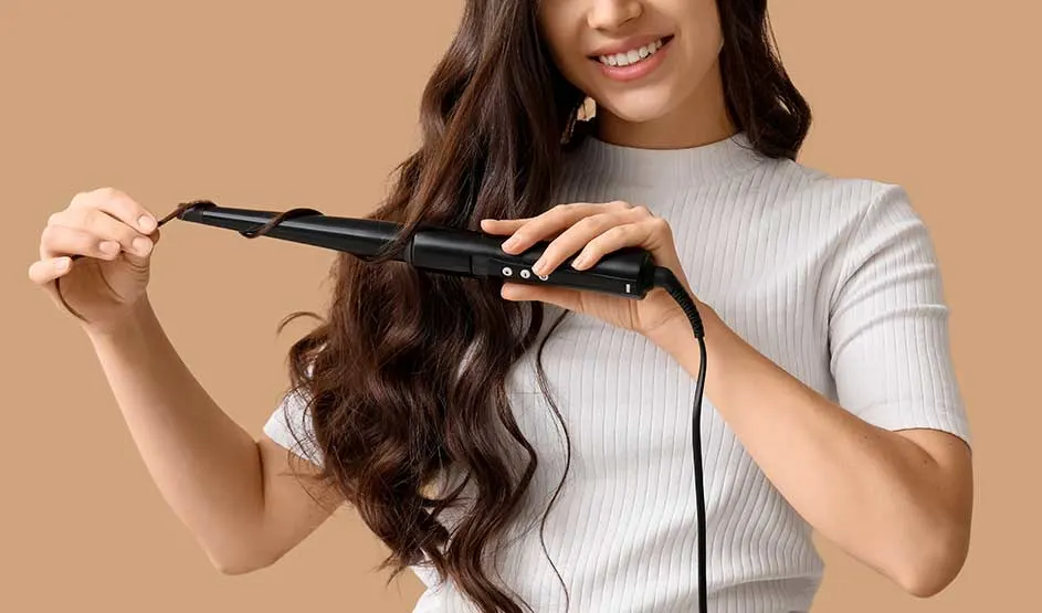 Person with long wavy hair using a black curling iron, dressed in a white ribbed top against a beige background.
