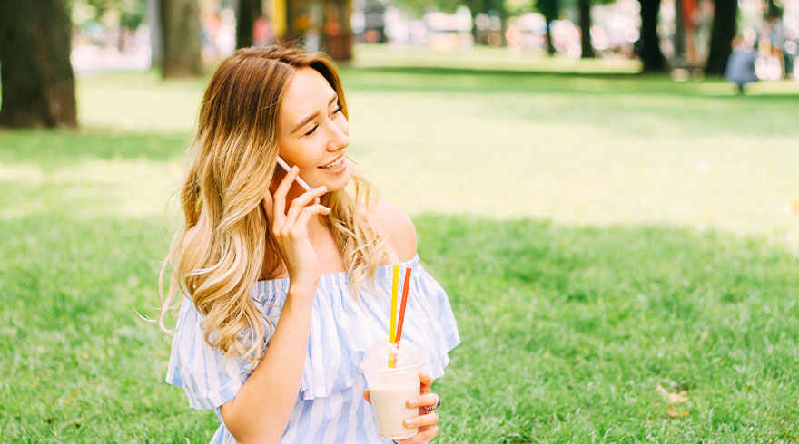 Vrouw met telefoon en drankje op gras in park. Ze draagt een gestreepte jurk met blote schouders. Bomen op achtergrond.
