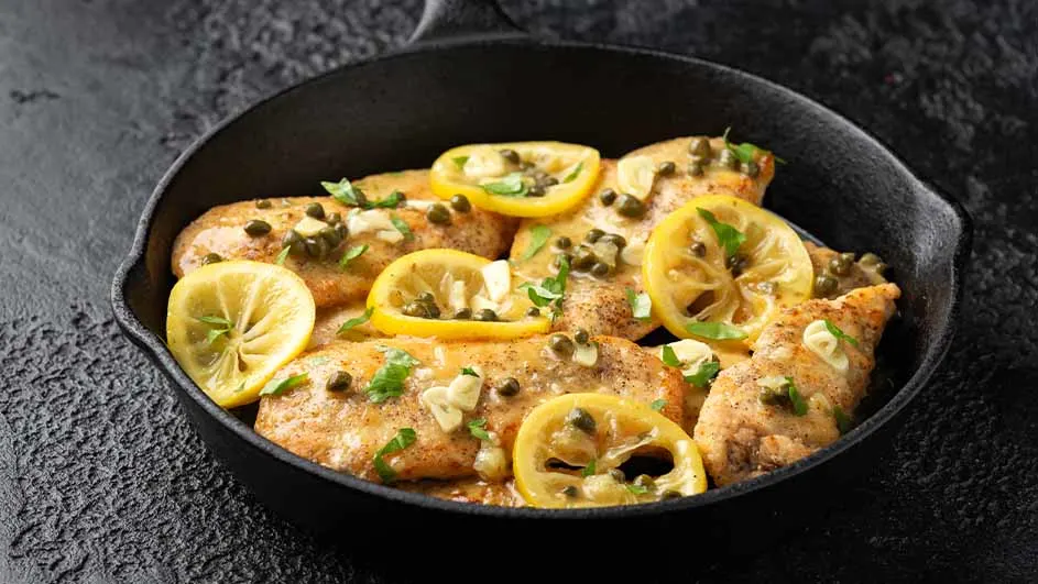 Gebakken kip met schijfjes citroen en kappertjes in een zwarte pan. De achtergrond is donker.