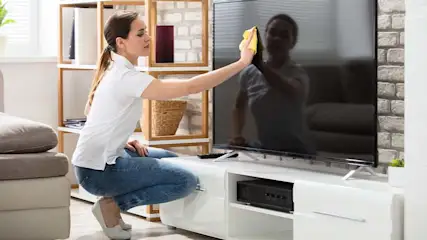 Een vrouw knielt voor een tv en maakt het scherm schoon met een gele doek. Achter haar staan een kast en een bank.