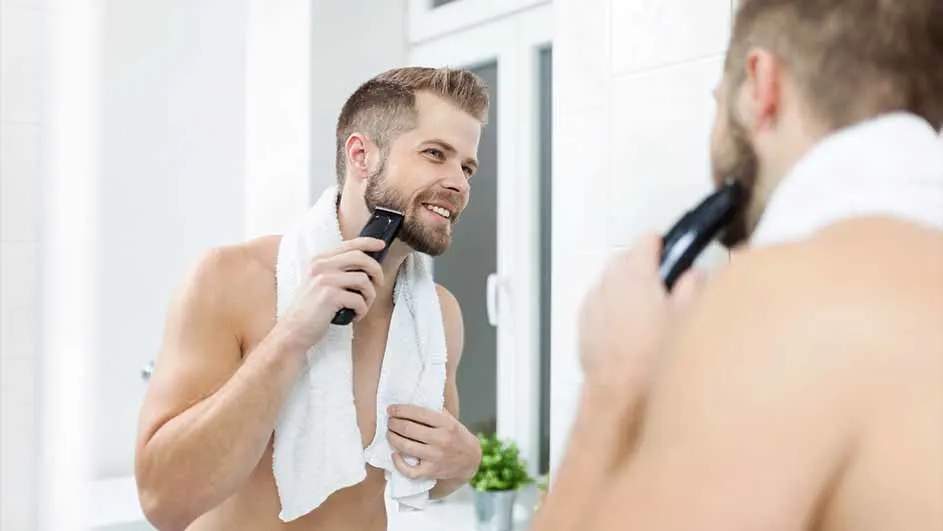 Een man scheert zijn baard in een witte badkamer met een zwarte trimmer, een witte handdoek om zijn nek.
