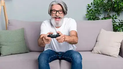 Een man met grijs haar en baard zit op een bank en speelt videogames met een zwarte gamecontroller. Op de achtergrond staan groene planten.