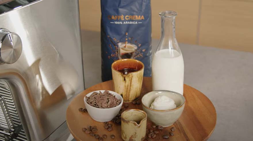 Een ronde houten plank toont koffiebonen, chocoladeschilfers, melk, room en een pak Arabica koffie.