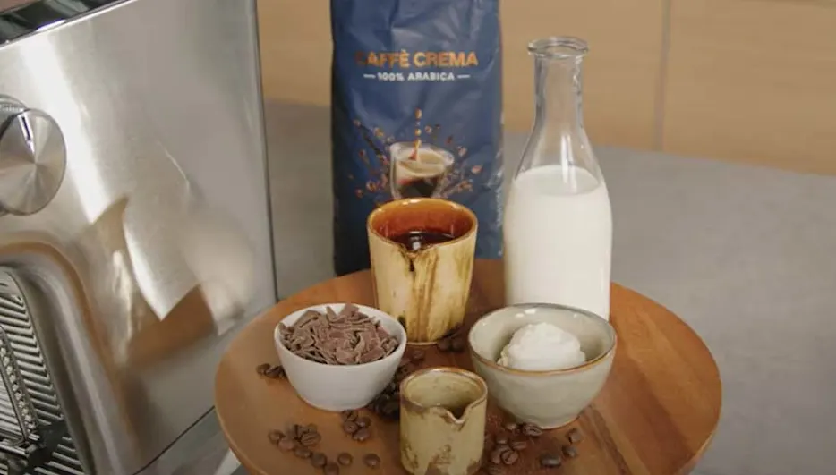 Een ronde houten plank toont koffiebonen, chocoladeschilfers, melk, room en een pak Arabica koffie.