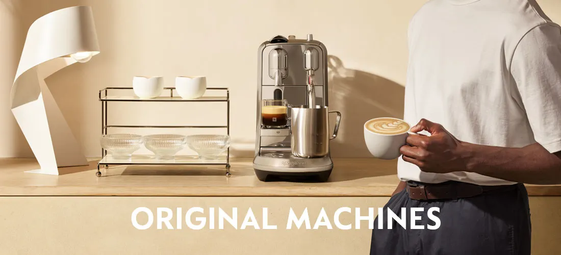 Koffiehoek met een Nespresso Original-machine op een houten blad; een persoon houdt een cappuccino vast, met lamp en rek met kopjes op de achtergrond.