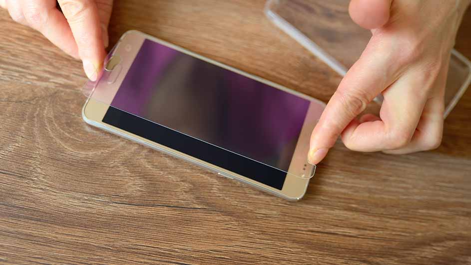 Twee handen plaatsen een schermbeschermer op een gouden smartphone, liggend op een houten tafel.