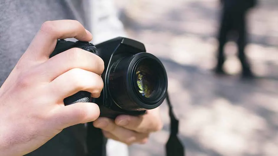 Een persoon houdt een zwarte camera vast met beide handen, met een vage figuur op de achtergrond.