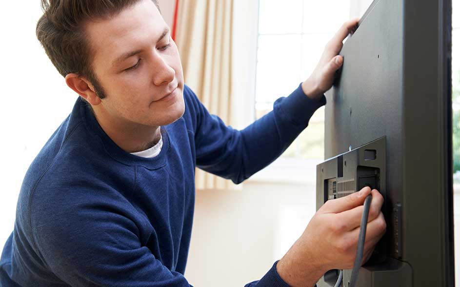 Een man in een blauwe trui sluit een kabel aan achter een flatscreen-tv; hij houdt het toestel vast in een lichte woonkamer bij het raam.
