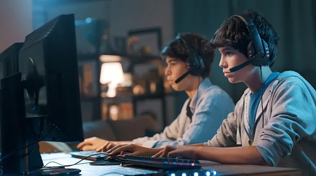 Two teenagers wearing headsets sit focused at large monitors, gaming together at the same desk in a cosy room.