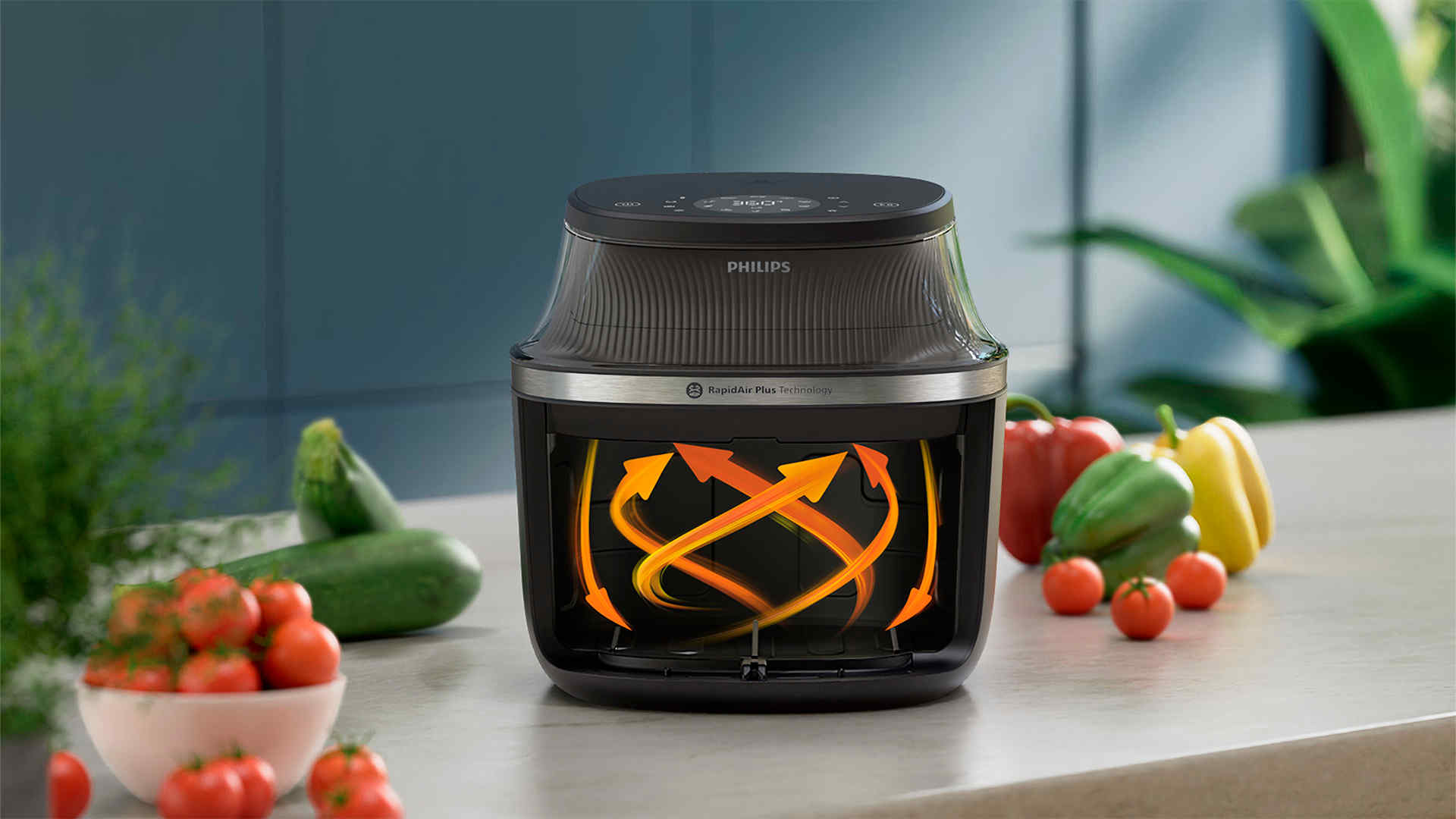 Een Philips airfryer met oranje pijlen binnenin, omringd door tomaten, courgettes en paprika's op een aanrecht.