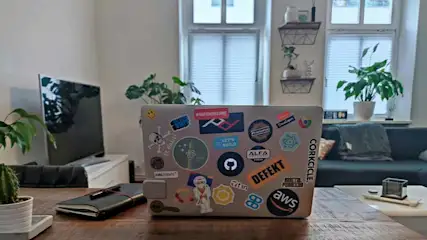 Een MacBook met allerlei stickers op de achterkant staat op een houten tafel in een woonkamer met veel planten, een bank en een televisie op de achtergrond.