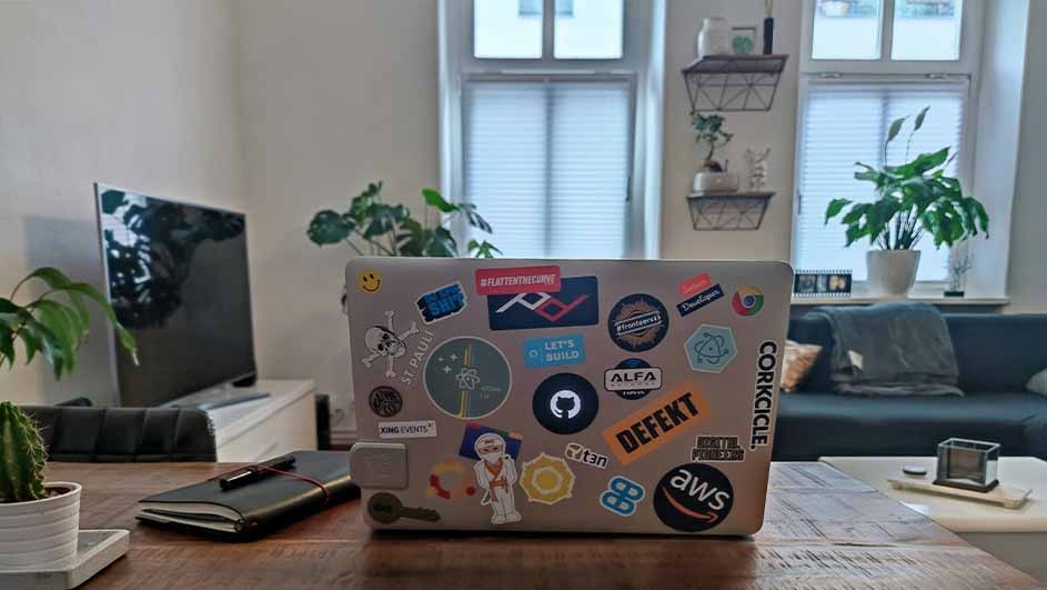Laptop with stickers on a wood table, notebook, cactus, TV, and couch visible in a room with windows.