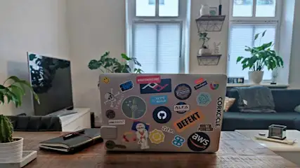 Laptop with stickers on a wood table, notebook, cactus, TV, and couch visible in a room with windows.