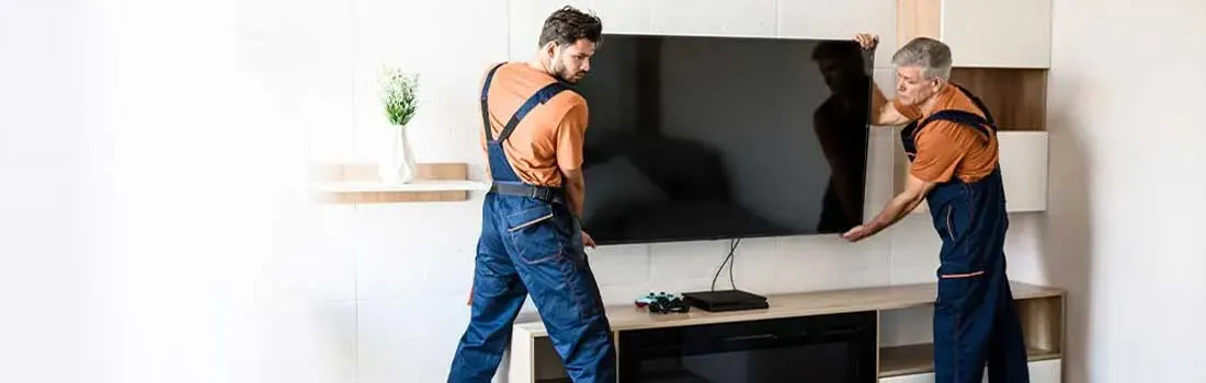 Two workers in blue overalls are installing a large, flat screen TV on a wooden entertainment center with a built-in fireplace.