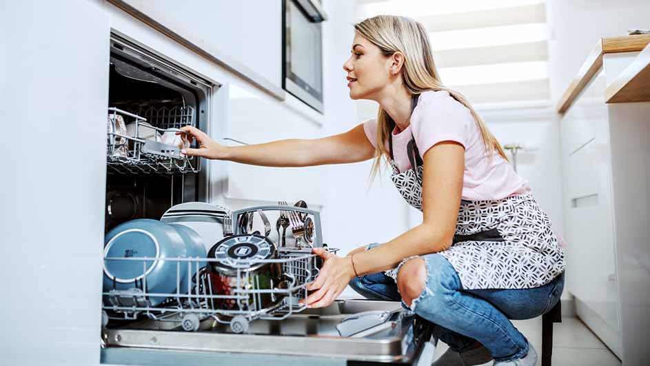 Vrouw laadt vaatwasser in, geknield met schort. Borden, bestek in rekken. Witte keukenachtergrond.