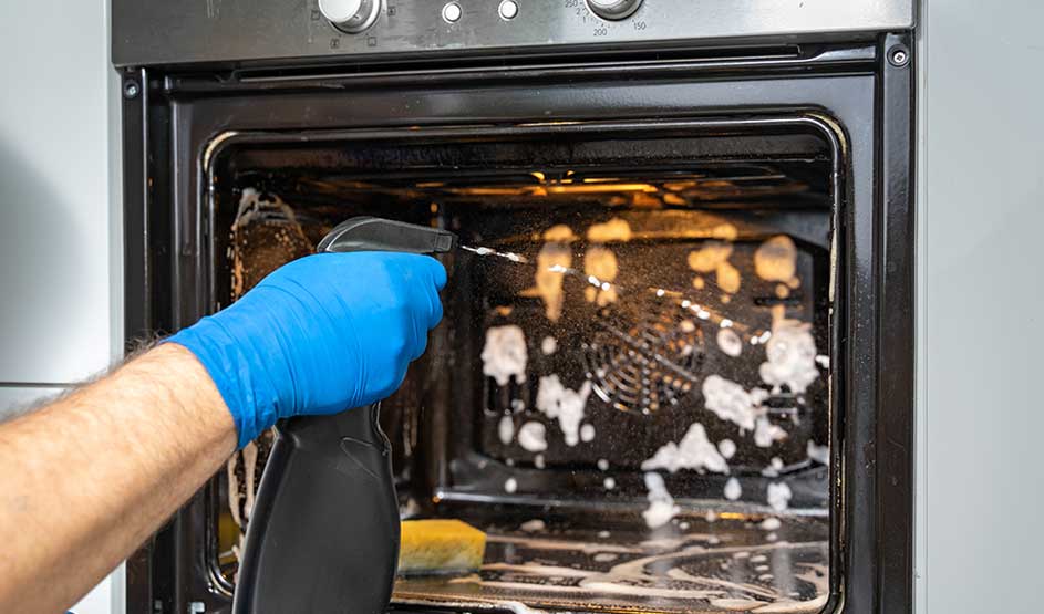Someone wearing a blue glove sprays cleaner into an open oven filled with foam and grime.