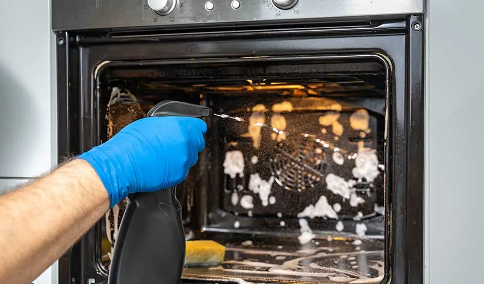 Iemand met een blauwe handschoen spuit schoonmaakmiddel in een open oven vol schuim en vuilresten.