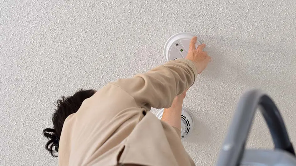 Iemand installeert een witte rookmelder aan een witte muur, staande op een grijze ladder.