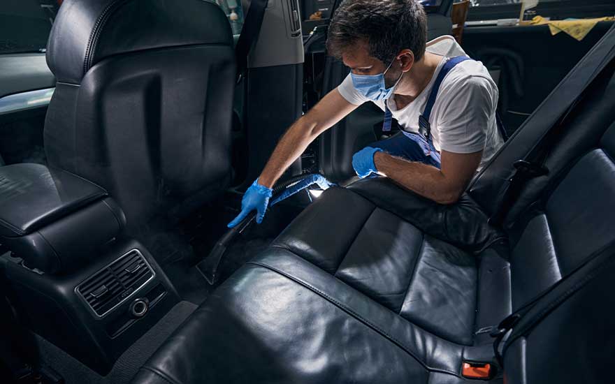 Een schoonmaker in overall en mondkapje gebruikt een stofzuiger om het vuil tussen de achterstoelen van een auto zorgvuldig te verwijderen.