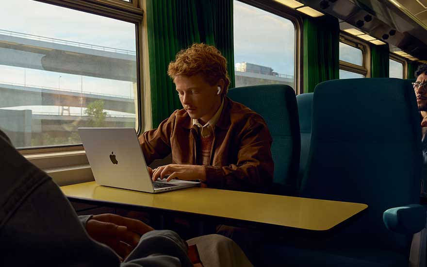 A young man works intently on his laptop at a train table, wearing wireless earbuds as the scenery outside the window passes quietly by.