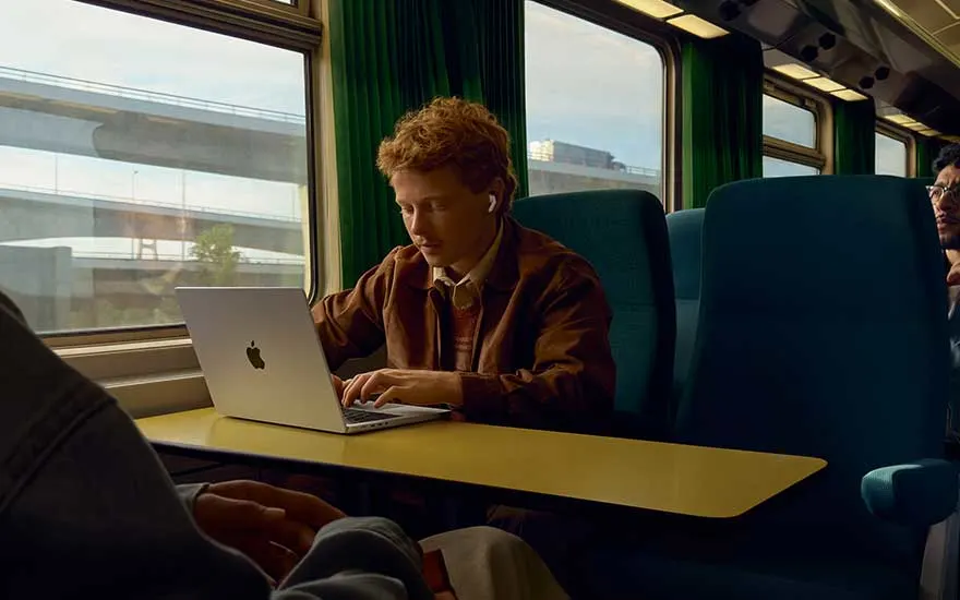 Jonge man werkt geconcentreerd op zijn laptop in een treincompartiment, met oordopjes in, terwijl het landschap buiten rustig langs het raam glijdt.