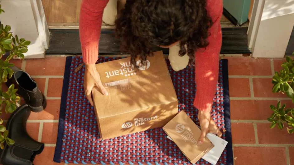Een persoon in een rode trui pakt een HP Instant Ink-pakket op van een blauw-rood vloerkleed bij een bakstenen ingang.