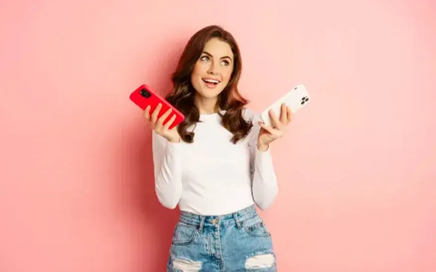 Een vrouw in een wit shirt en jeans houdt een rode en een witte telefoon vast voor een roze achtergrond.