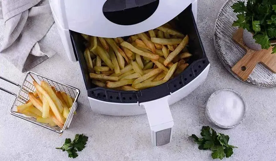 White air fryer with a drawer full of golden chips (fries), fresh parsley and salt alongside. Brand is not visible on the appliance.