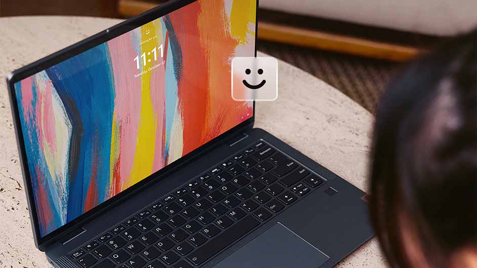 Laptop met kleurrijk scherm en emoticon, staand op een tafel. Een persoon zit rechts van de laptop.