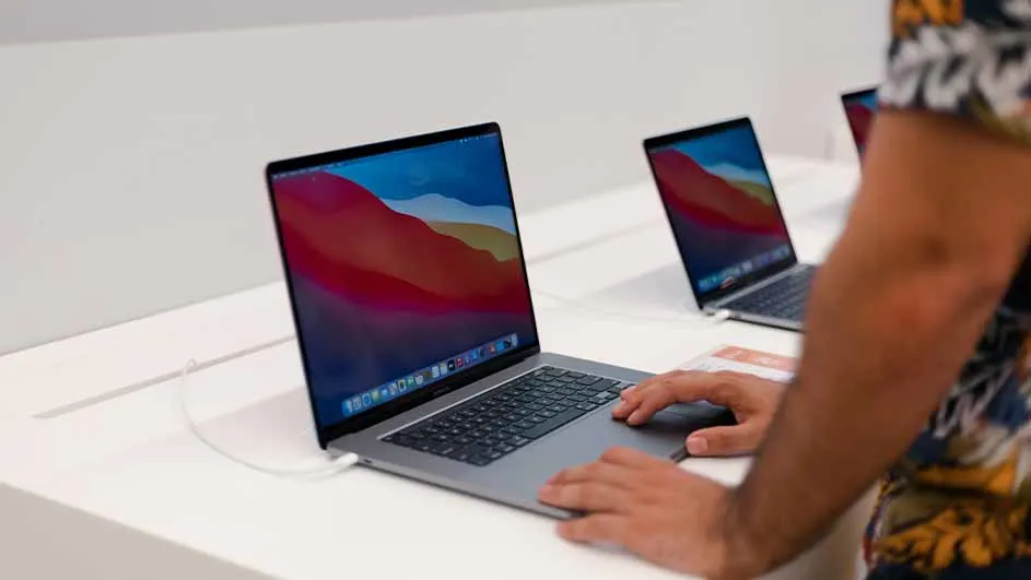 Een persoon gebruikt een grijze laptop op een witte tafel. Er staan andere laptops op de achtergrond, allemaal met schermen aan.