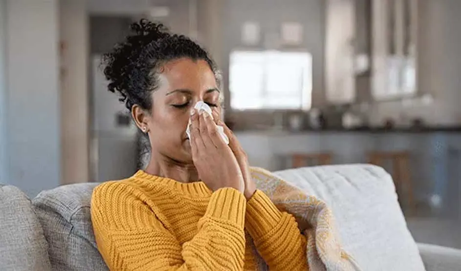 A person sits on a sofa in a cosy living room, eyes closed, gently touching their nose with a soft tissue held tussen beide handen.