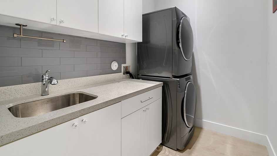 A laundry room with a sink, countertop, and white cabinets under a gray brick backsplash. Next to it, a stacked gray washer and dryer.