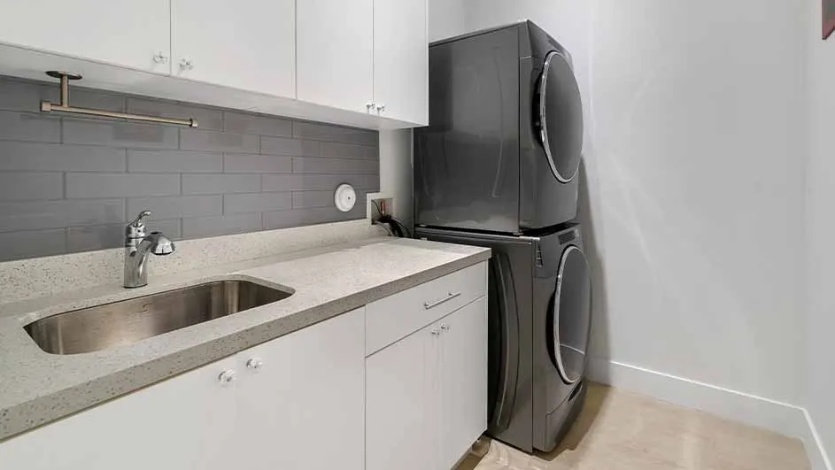A laundry room with a sink, countertop, and white cabinets under a gray brick backsplash. Next to it, a stacked gray washer and dryer.