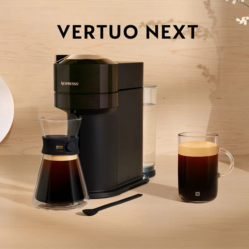 Een zwarte Nespresso-machine naast een glazen beker met koffie en een lepel op een houten tafel.